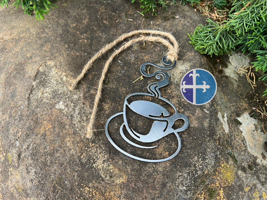 Steaming steel coffee cup ornament with rustic twine, lying on a textured stone surface surrounded by greenery.