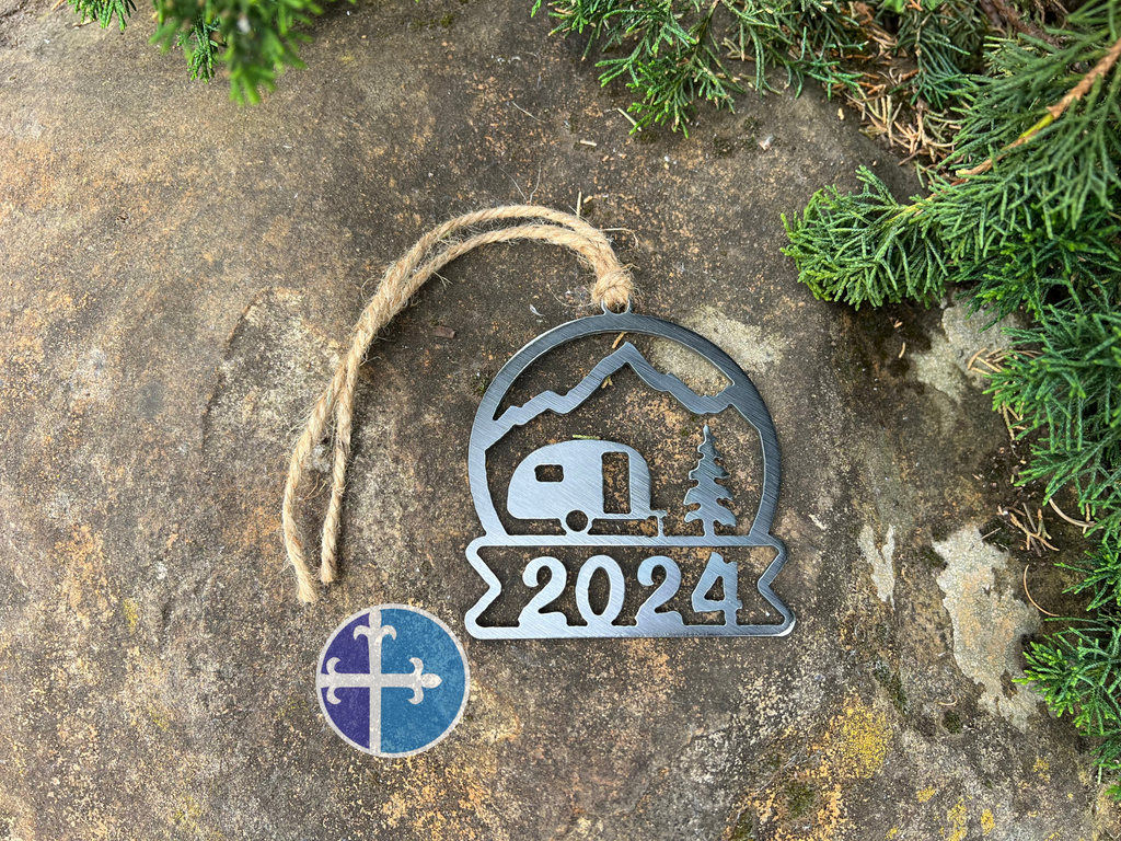 A metal 2024 camper ornament with a mountain and tree design, hanging by a rustic twine on a natural background.