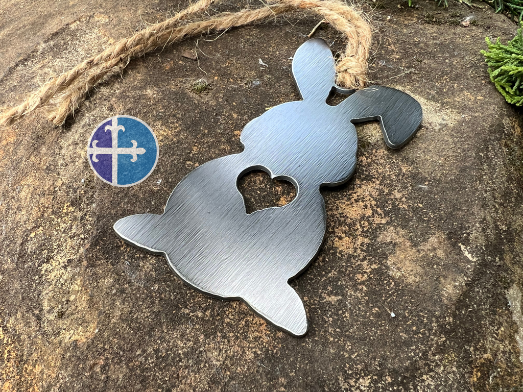 Close-up of a rustic steel bunny ornament with a heart cutout in the center, hanging from a jute twine on a textured stone background.